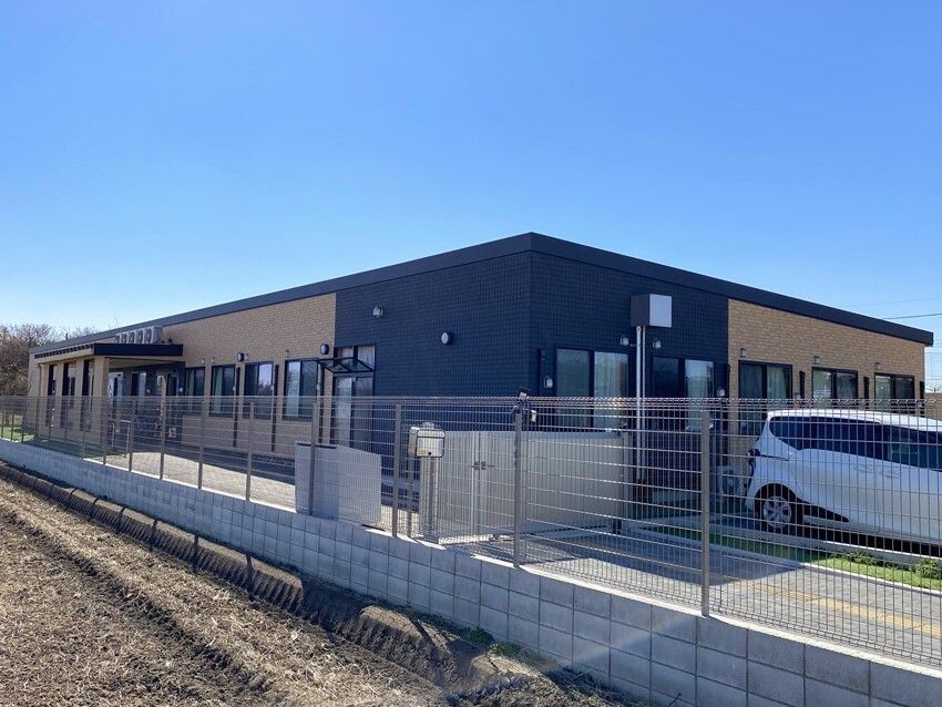 日当たりのいい場所に建っている平屋建ての建物の周りには畑があり、駐車場には車が止まっている。