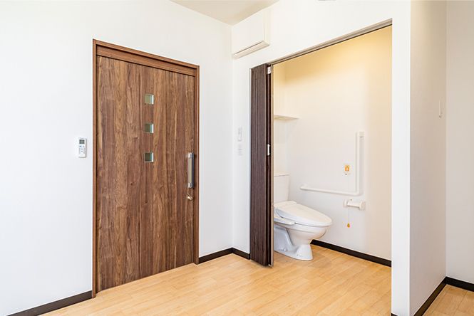 オレンジの呼び出しボタンやエル字型の手すりが設置された広いトイレがフローリングの居室に付き、すぐそばに出入り口がある。