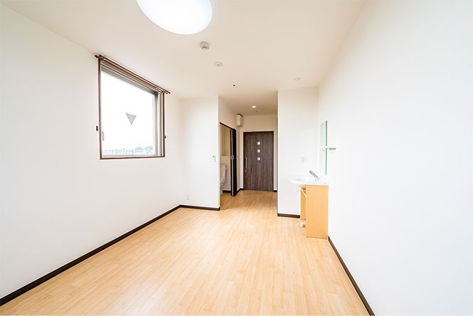 大きな窓が左、洗面所が右にあり、左奥にはトイレが設置されていて、家具などは置かれていない広々としたフローリングの部屋。