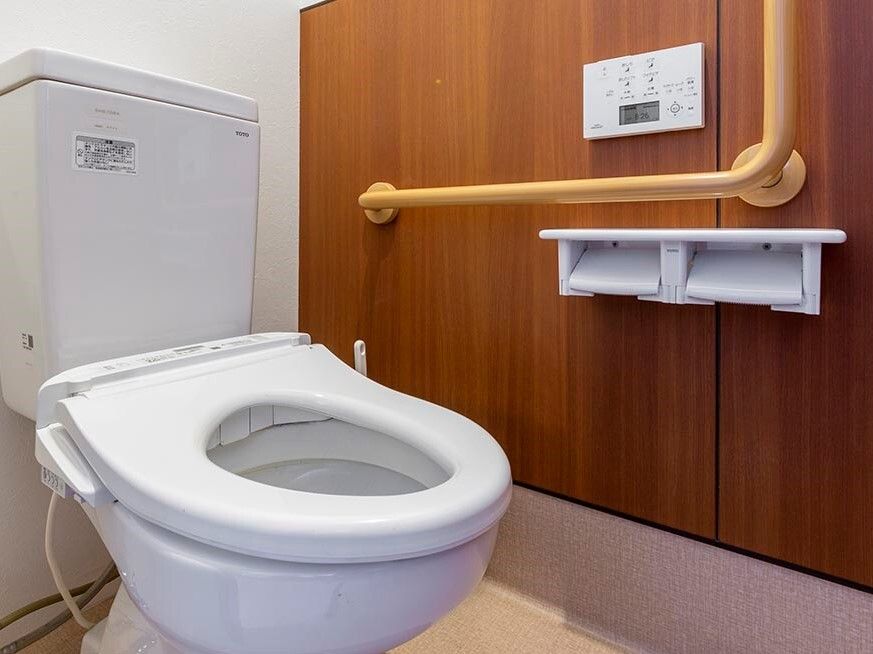 エル字型の手すりや操作パネル、トイレットペーパーホルダーが壁に付いたトイレ。便座にふたはなく、シンプルなデザインになっている。