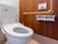 エル字型の手すりや操作パネル、トイレットペーパーホルダーが壁に設置されたトイレ。便座にはふたがなく、シンプルなデザイン。