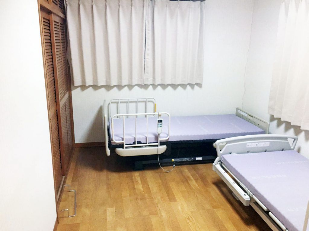 白いカーテンが設置された窓が二つ付いたフローリングの居室。介護用ベッドが二つあり、大きなクローゼットも付いている。