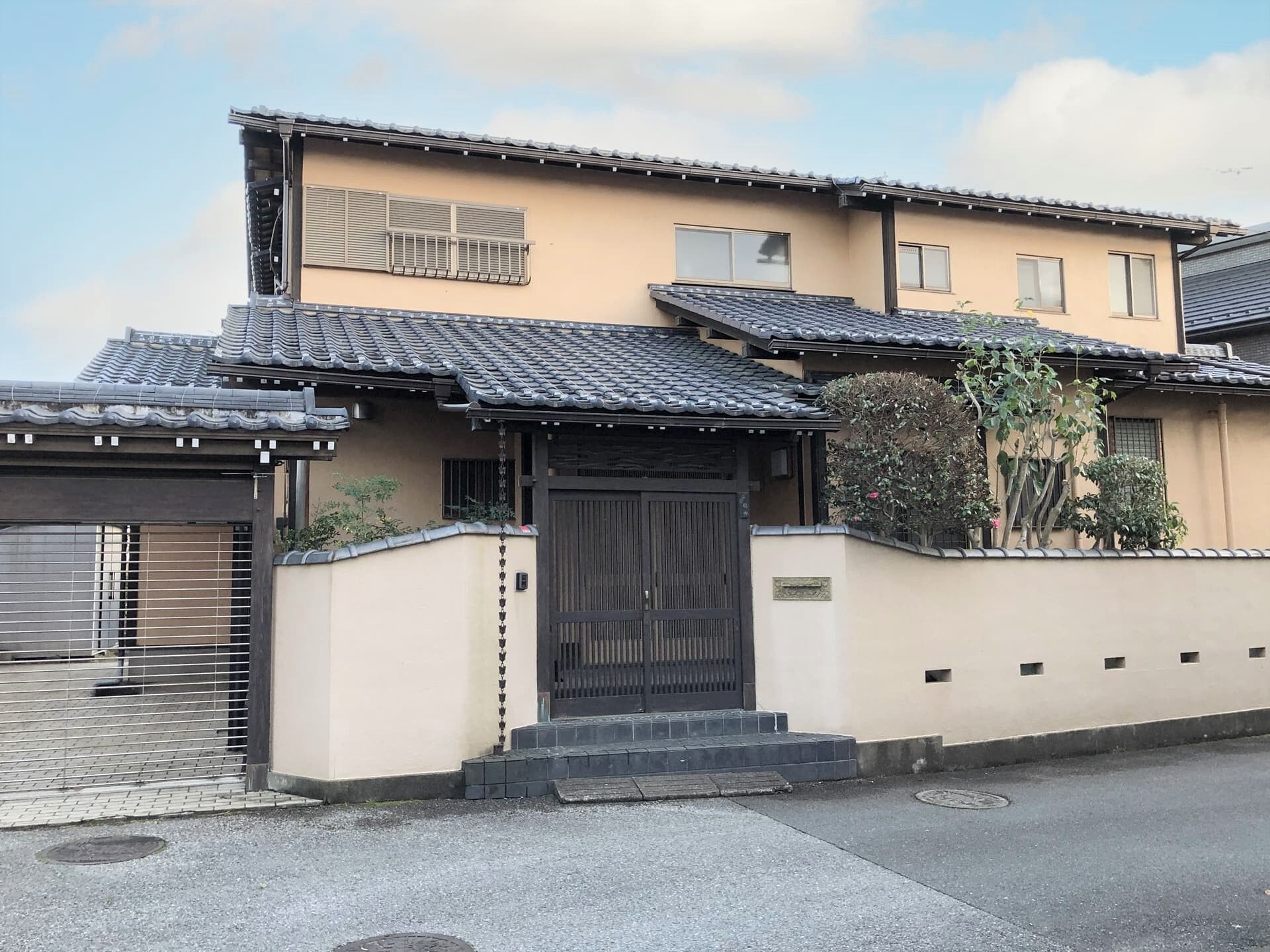 2階建ての建物は和風でグレーの瓦屋根が付いている。建物の周りには木が植えられ、玄関の前には階段があり、駐車スペースもある。