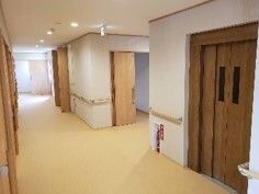 緑の多い屋外で笑顔で過ごしている眼鏡姿の高齢女性と付き添いのブルーのポロシャツを着た若い女性スタッフ。高齢女性は白髪でショートカット。