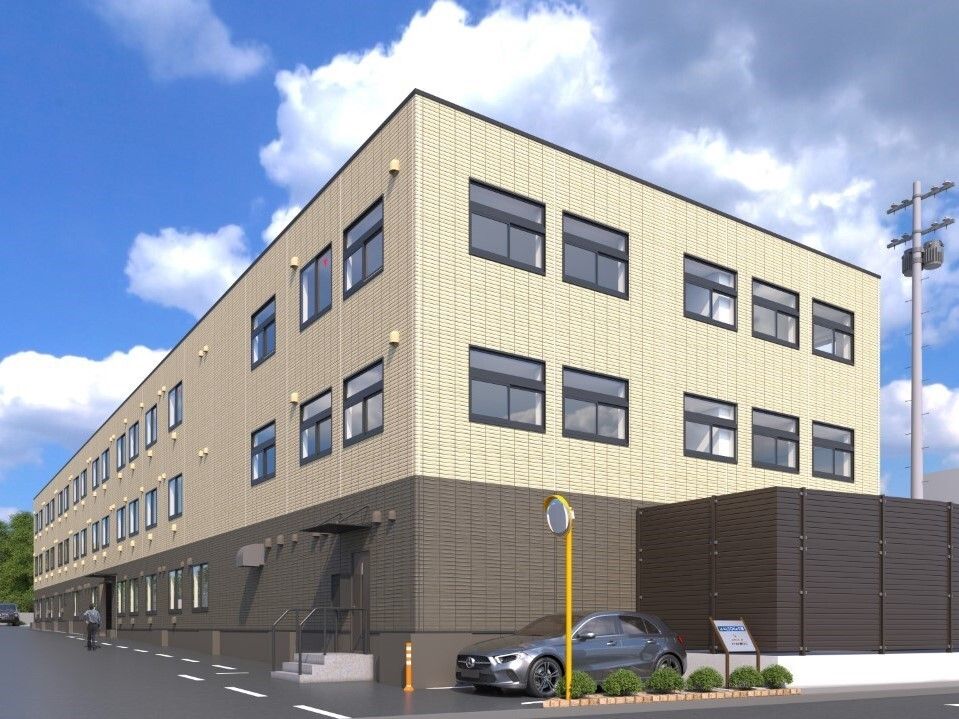 1階部分がブラウン、2階と3階がベージュの3階建ての建物は横に長く、小さいが駐車スペースもある。上には青空が広がっている。