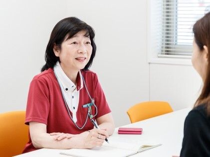 女性スタッフがペンを持って紙にメモを取りながら髪の長い女性の話を笑みを浮かべて聞いている。