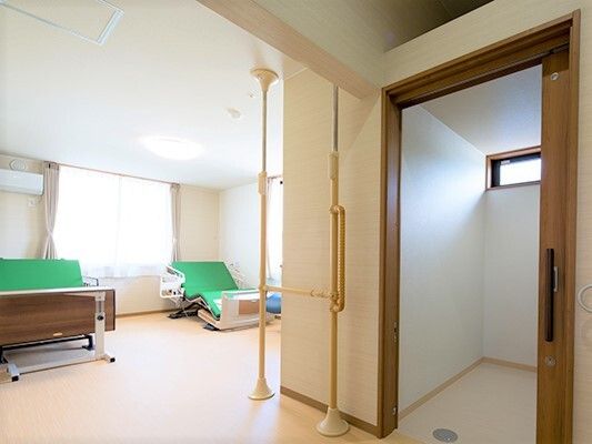 トイレ付きの二人部屋。動かすことができる大きな手すりが壁際に付き、緑の介護ベッドが２台並んでいて、壁にはエアコンもある。