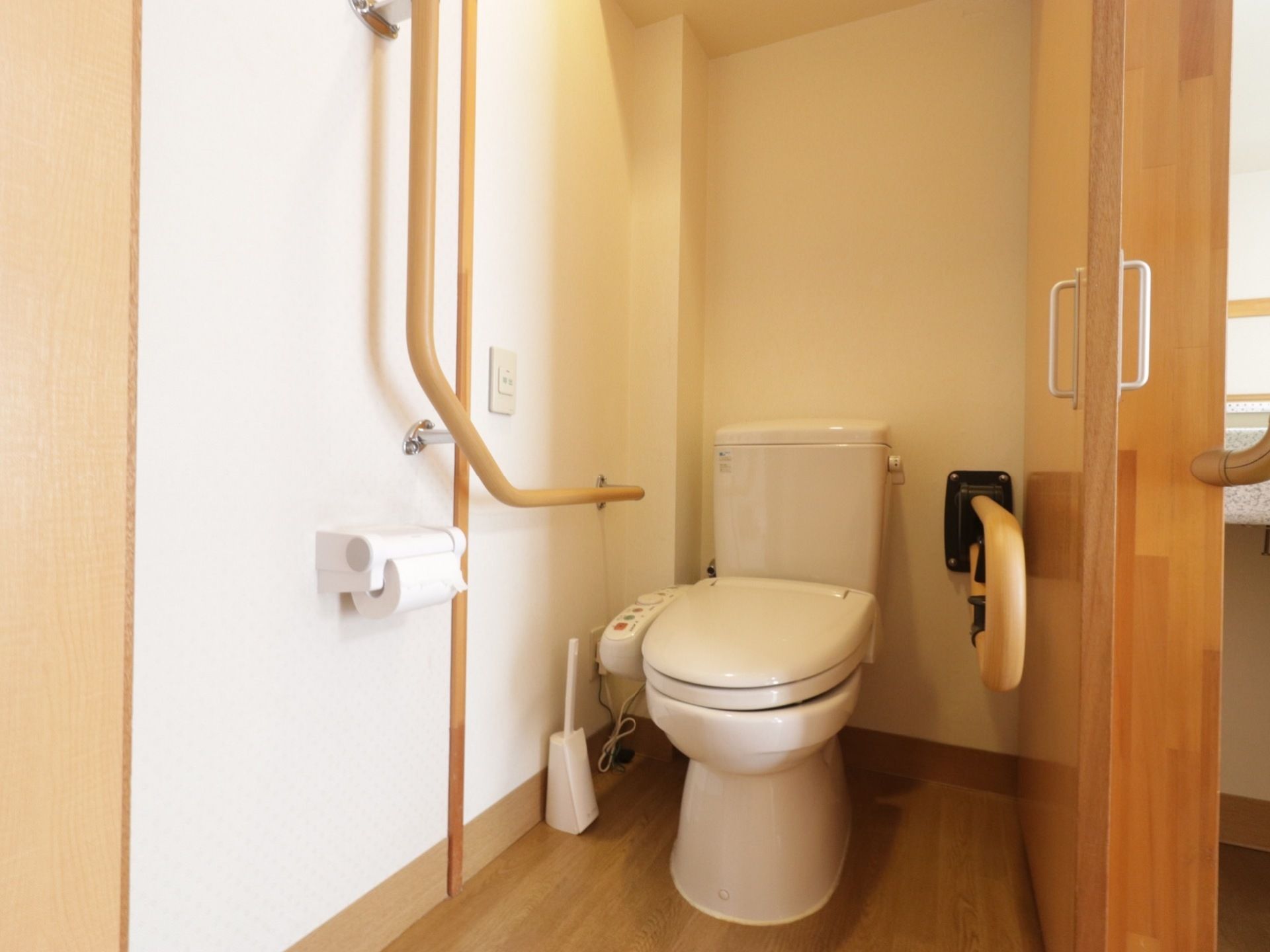 白とライトブラウンで統一された明るいデザインのトイレである。左側の壁にはL字型の手すりがあり、すぐ側に呼出ボタンがある。