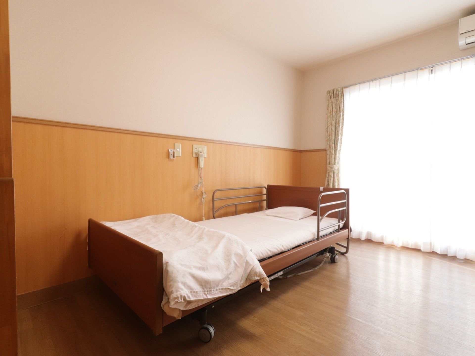 居室の壁は、白とベージュ色のツートンカラーになっている。壁際には大きな電動ベッドがあり、布団には真っ白なカバーが掛けてある。
