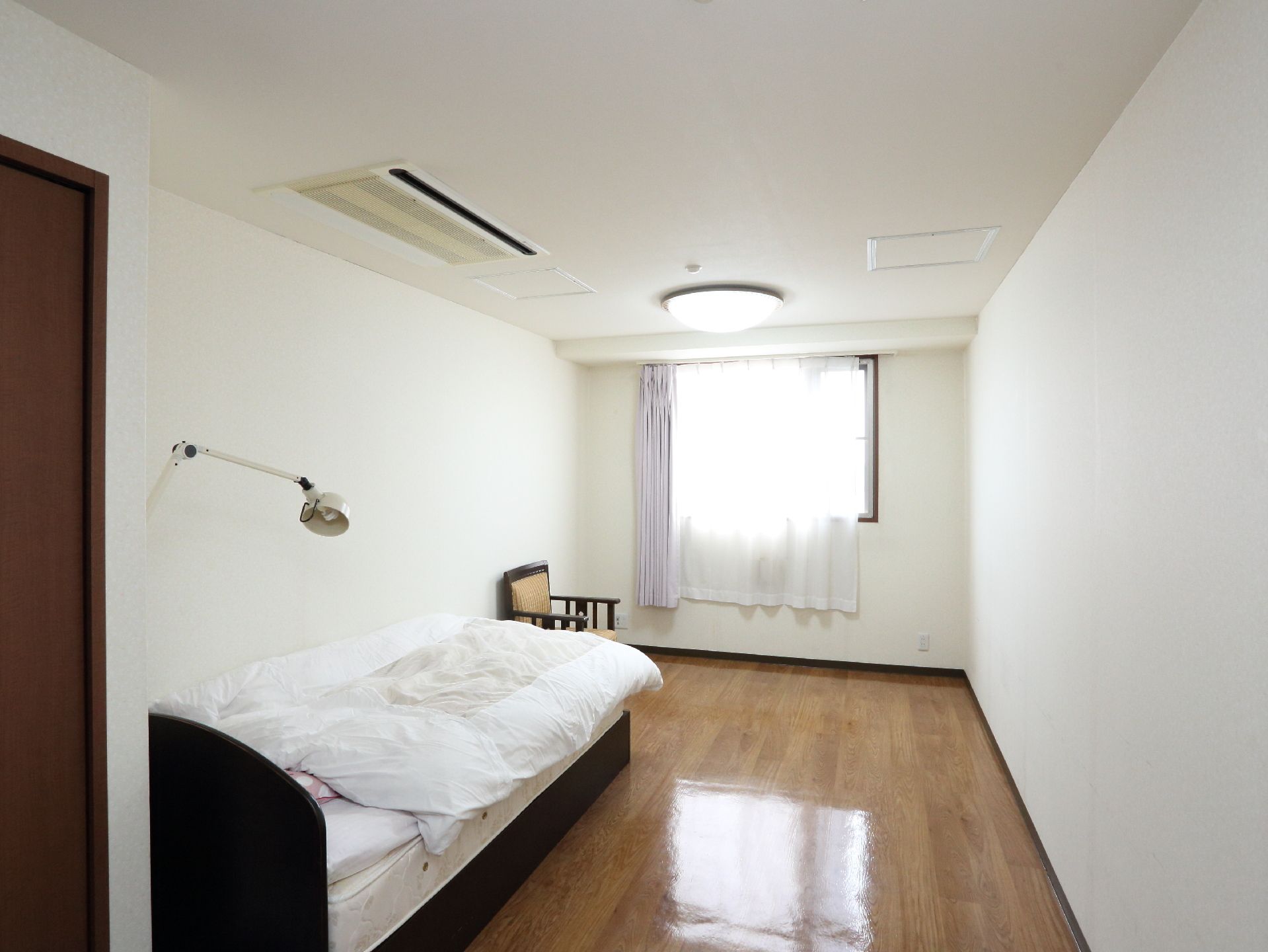 大きな窓が付いた明るいフローリングの部屋には寝具付きのベッドやエアコン、大きなクローゼット、グレーの一人用の椅子が設置されている。