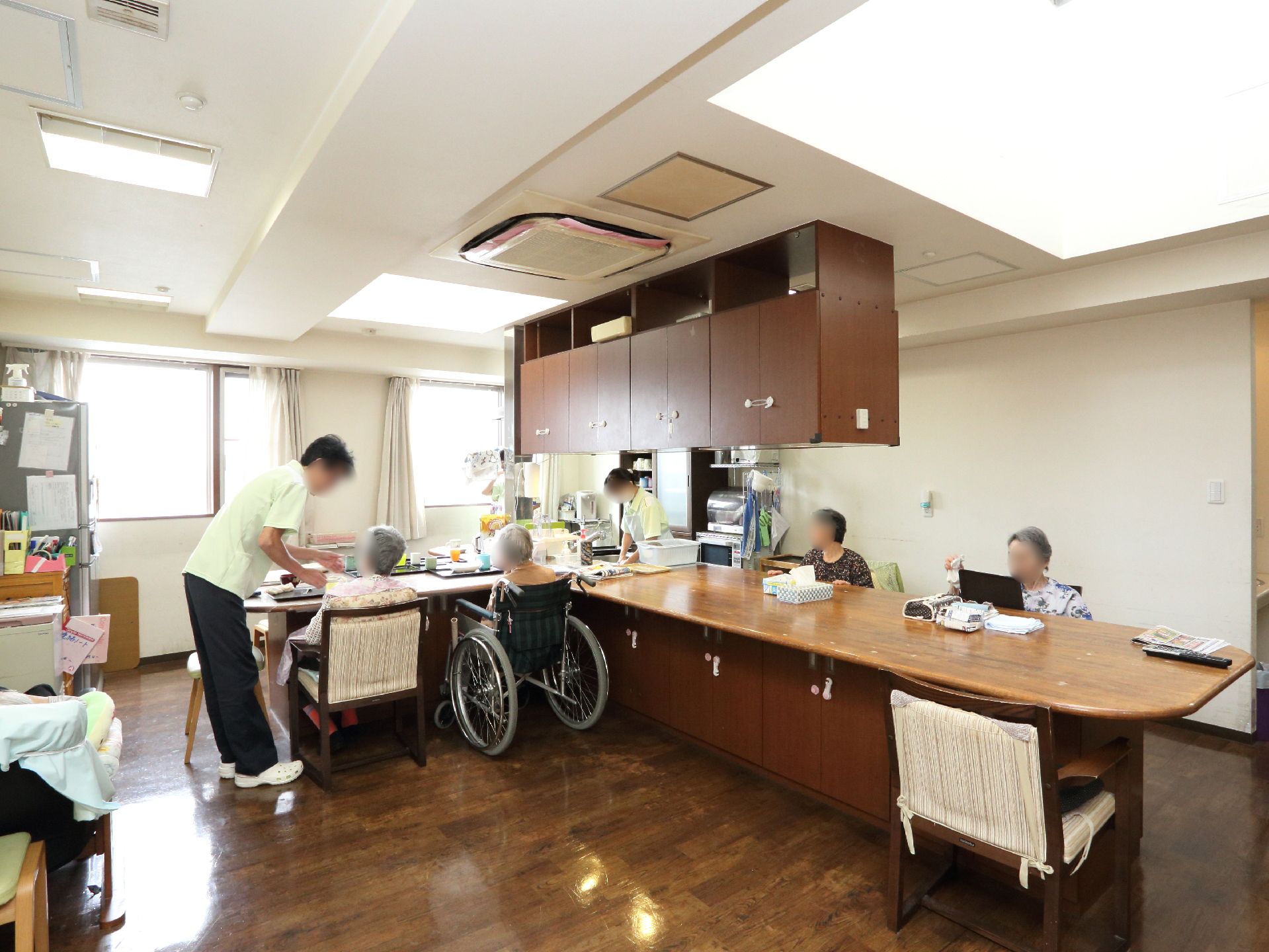 共有ルームには車いすに乗っている入居者様や、そのお世話をしているスタッフがいる。天井にはいくつかの照明がある。