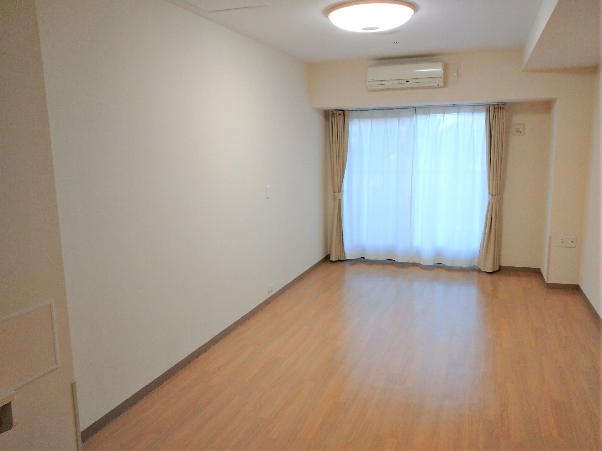 エアコンや照明が付いたシンプルなフローリングの部屋。大きな窓にはカーテンとレースカーテンが付き、壁は白く清潔感がある。