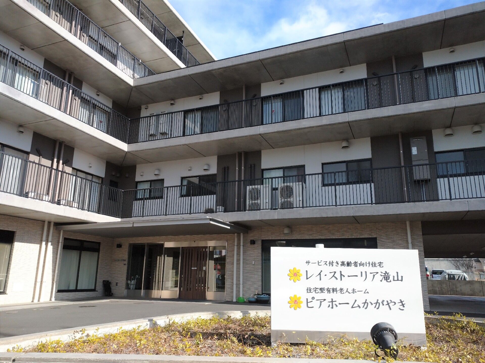 白とグレーの建物は5階建てで、玄関がある部分は一部3階建てとなっている。各フロアにベランダが付き、玄関には屋根があり、敷地が広い。