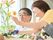 たくさんの花が置かれたテーブル。フラワーアレンジメントを楽しんでいる眼鏡をかけた高齢男性のサポートをエプロン姿の女性スタッフが行っている。