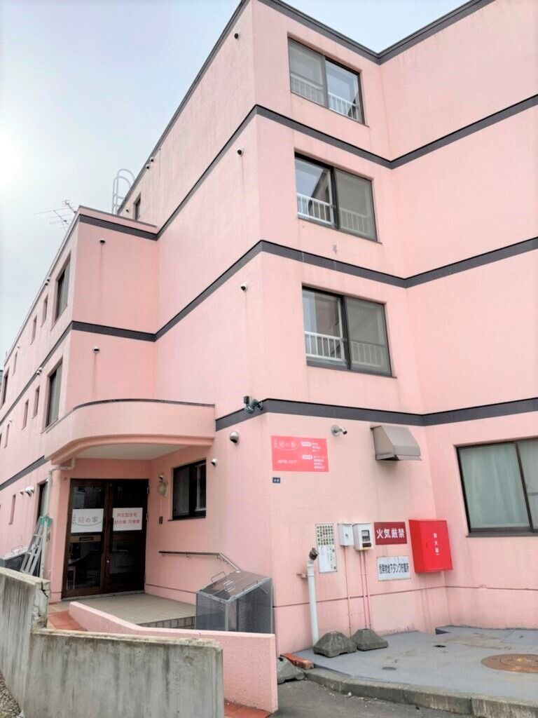 手すり付きのスロープが玄関に付いたピンクの建物。4階建てで玄関には屋根があり、建物を囲むように背の低い壁も作られている。