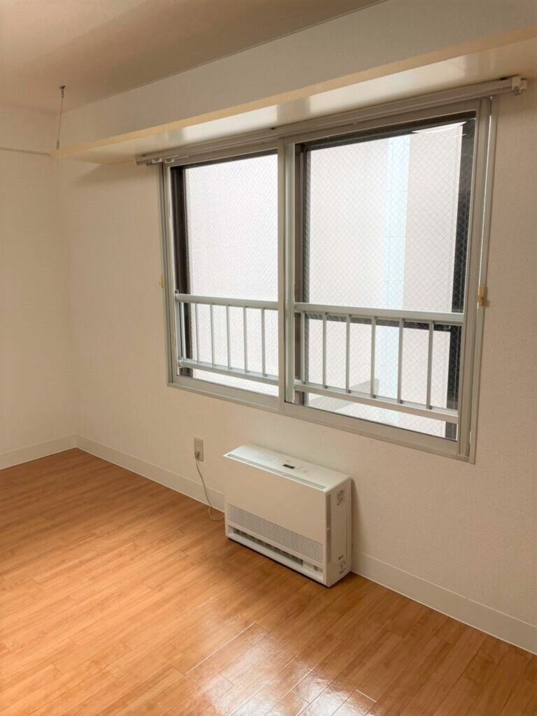 柵がついた窓にはカーテンが付いていない。家具や家電が置かれていないシンプルなフローリングの部屋には暖房器具のみ置かれている。