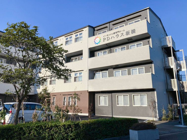 ライトグレー系の建物の外壁にはホーム名が書かれている。周囲にはたくさんの木が植えられ、ベランダもある。