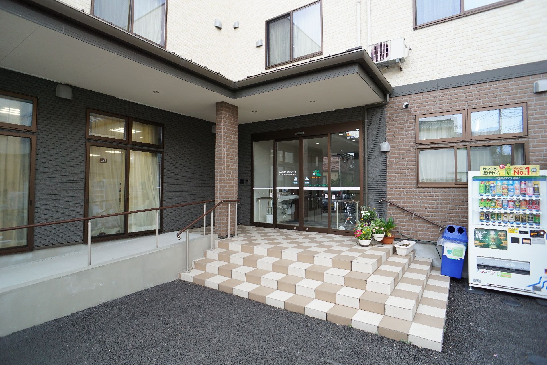 屋外で散策を楽しんでいる車いすの高齢女性と付き添いの若い女性スタッフ。二人は日当たりのいい場所で笑顔で過ごしている。