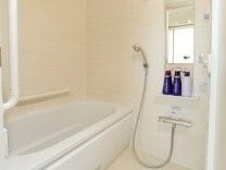 シャワーや手すりが設置された一人用の浴槽がある白を基調とした浴室。シャンプーなども置かれている。