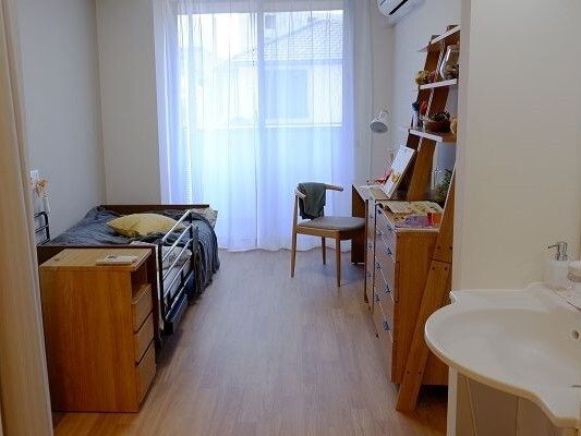 レースカーテンが付いた大きな窓があるフローリングの部屋にはエアコンが付き、寝具付きのベッドが置かれている。