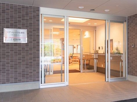 大きな3階建ての建物は横に長く、各フロアにベランダがあり、周辺には草木が植えられ、他に高い建物はない。玄関先で2人立ち話をしている人がいる。