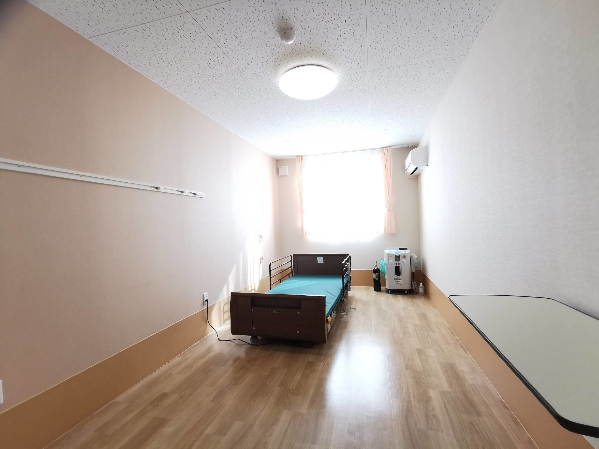 窓が大きく日当たりのいいフローリングの部屋。介護ベッドやエアコンが付き、医療機器も置かれている。壁にはピクチャーレールが設置されている。