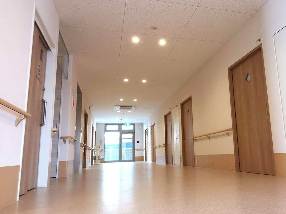両サイドに手すりや居室のドアが並んだまっすぐな廊下。突き当りにはガラスのドアがあり、日当たりがよく、居室のドアには丸い小さな窓が付いている。