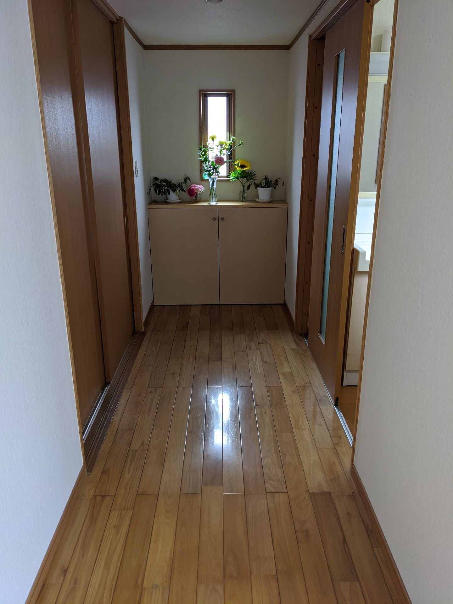きれいに磨かれたフローリングの廊下はまっすぐで右側には居室のドアらしきものがあり、正面の収納棚の上にはヒマワリなどの花が飾られている。