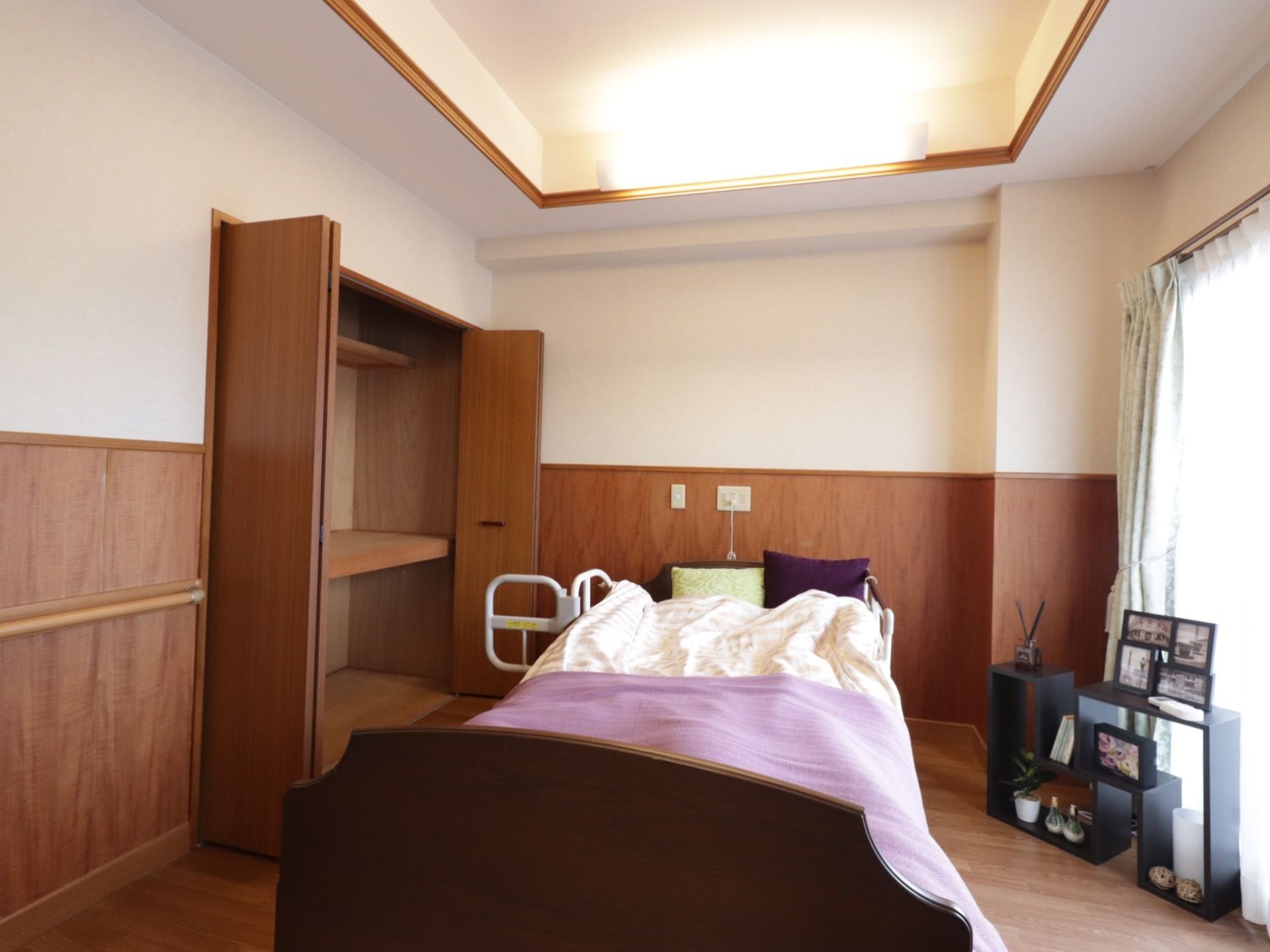 居室の中央に大きな電動ベッドがあり、薄紫色と白の布団カバーが掛けてある。左横に、折れ戸が付いた広い収納場所がある。