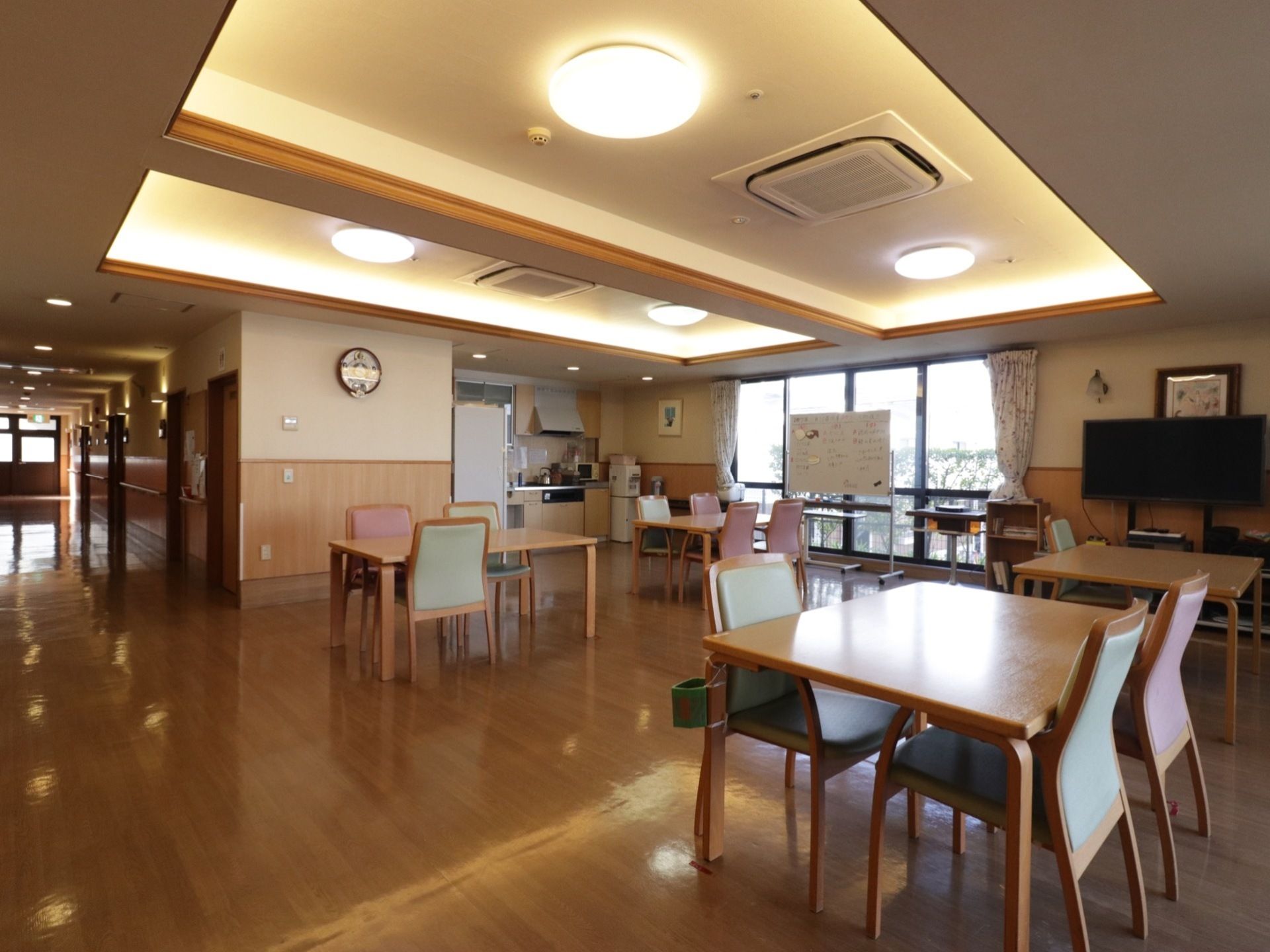 食堂の窓際には、大型テレビ、収納ボックス、ホワイトボード等が置いてある。大きな窓の左右には花柄のカーテンがまとめてある。