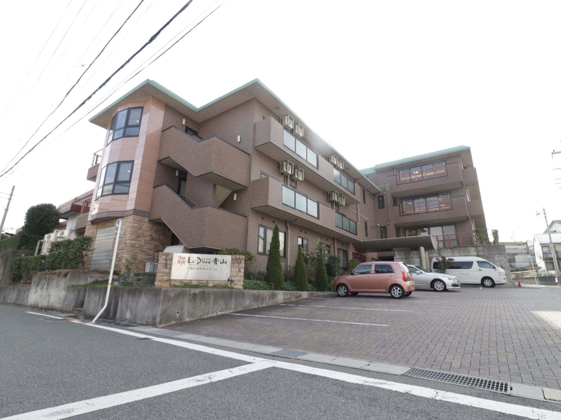 ３階建ての施設の右側に、広々とした駐車スペースがあり、軽自動車など数台の車が置いてある。道路脇には、大きな看板がある。