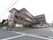 ３階建ての施設の右側に、広々とした駐車スペースがあり、軽自動車など数台の車が置いてある。道路脇には、大きな看板がある。