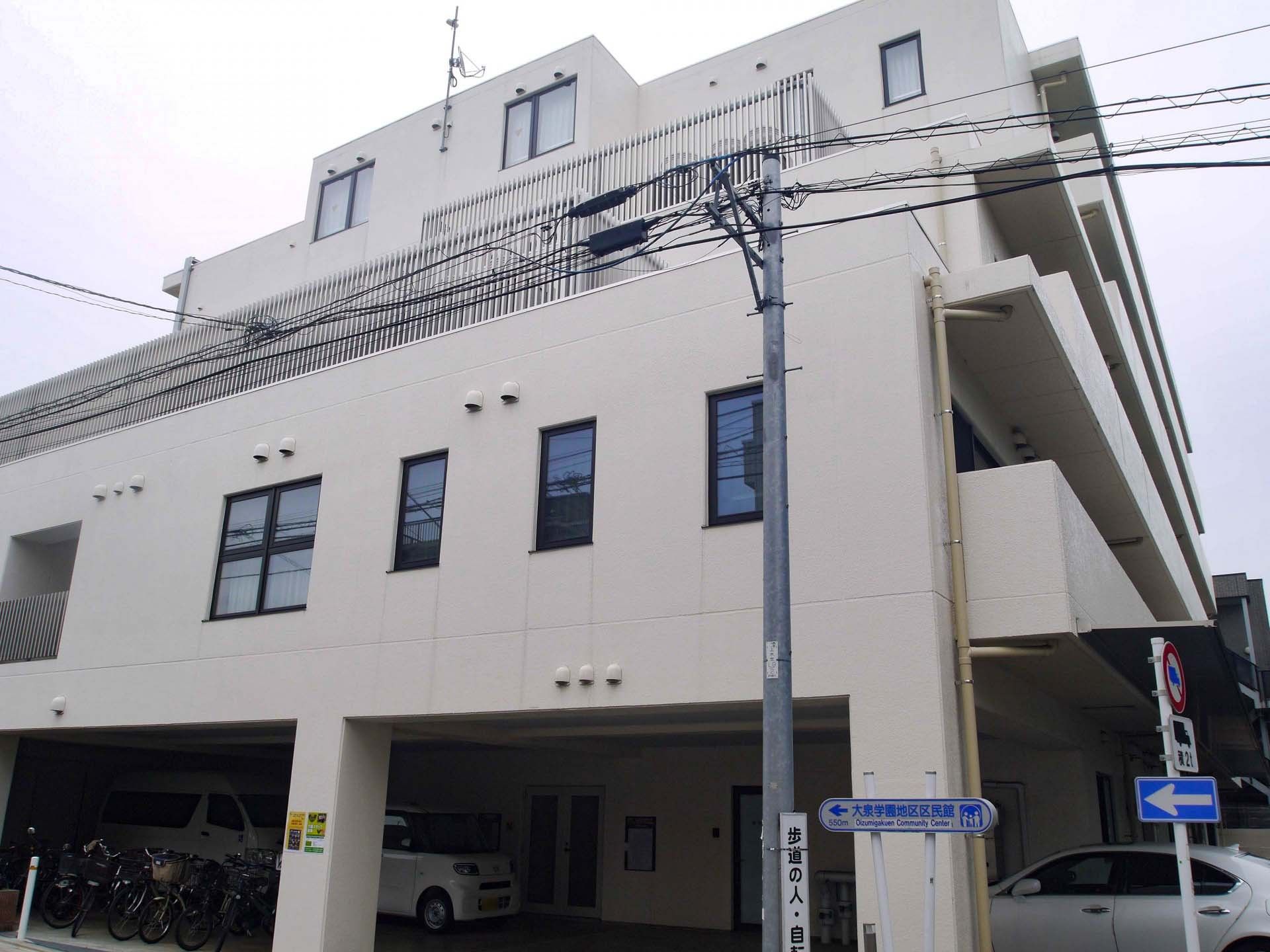たくさんの木が周りに植えられた白い5階建ての建物は道路に面していて、横断歩道が目の前にある。