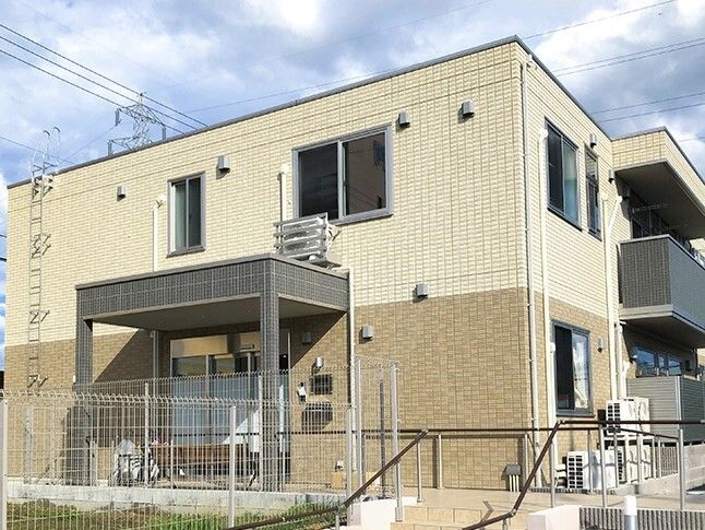 ベージュ系の２階建ての建物は日当たりがよく、玄関には手すり付きのスロープと屋根が付いている。