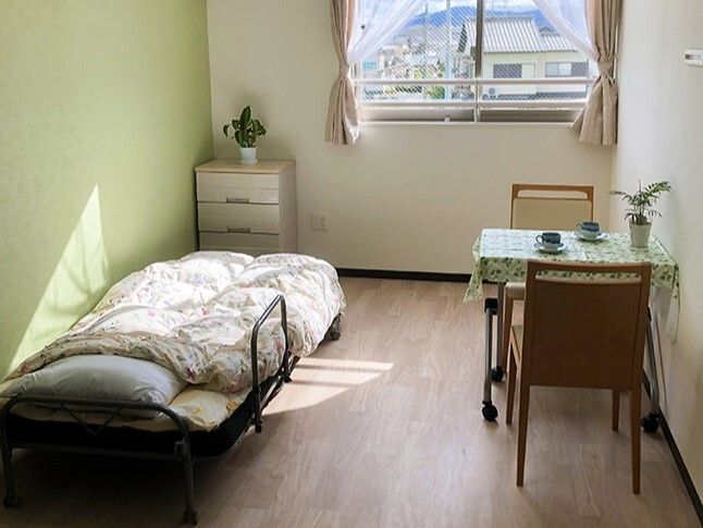 カーテン付きの窓があるフローリングの部屋には寝具付きのベッドやテーブルセットが置かれている。
