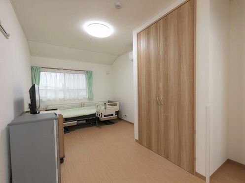 寝具付きのベッドが設置された居室には大型の収納スペースがあり、タンスの上には薄型のテレビがあり、大きな窓には緑のカーテンが付いている。