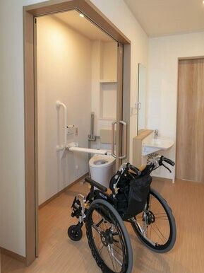 黒い車いすがトイレの出入り口に置かれている。トイレには背もたれや手すり、前方にバーが付き、ふたはないデザイン。