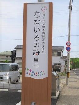 住宅地の中にある住まいで周辺には緑も多く残っている。住宅名が縦書きに書かれた看板の後ろには駐車場のような場所も見える。