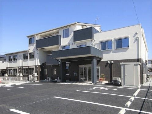 3階建ての建物は窓が多く、玄関部分には屋根と自動ドアが付いている。1階部分のみグレーで、上のフロアは白く、青い空が広がっている。