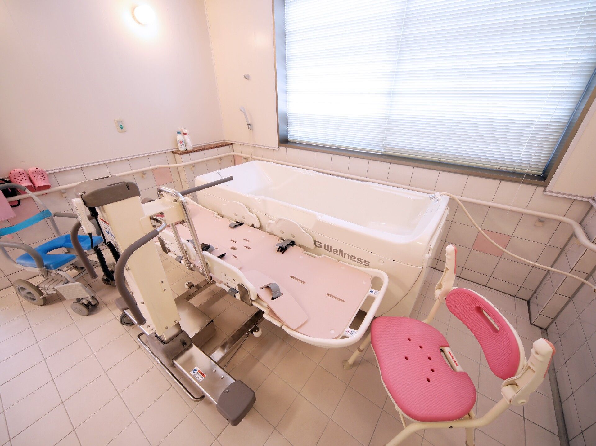 白い浴槽タイプの機械のそばにはストレッチャータイプのリフトがあり、シャワーチェアや浴室用車いすもある。