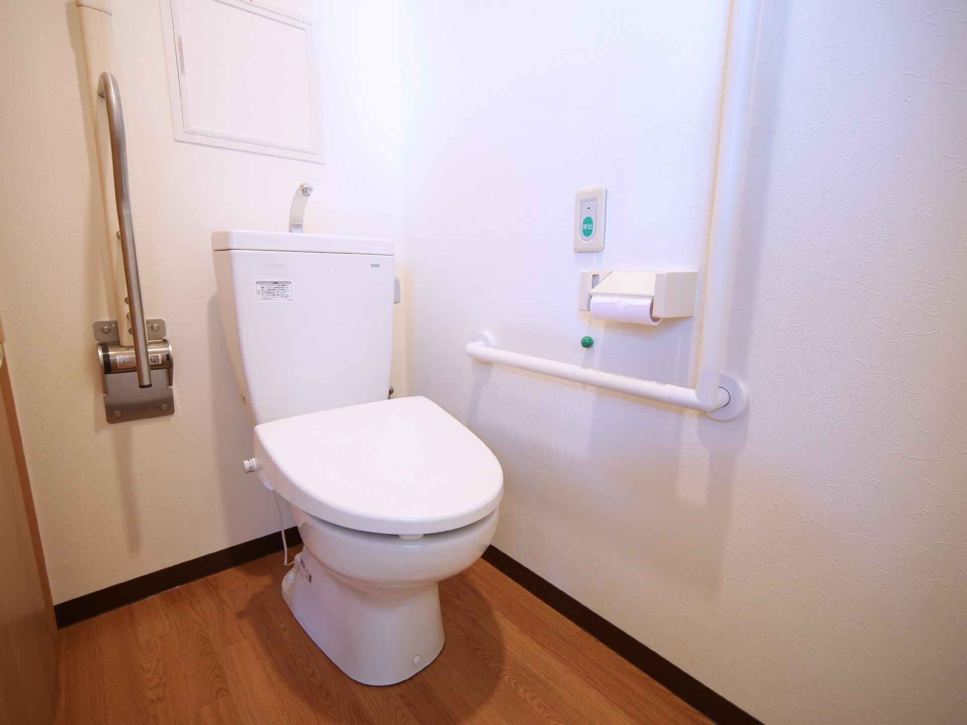 エル字型の手すりやグリーンの呼び出しボタン、トイレットペーパーホルダーが壁に付き、可動式の手すりも付いている。