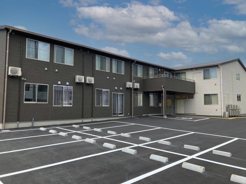 広い駐車場があり、ダークグレーの外壁の２階建ての建物は日当たり良好。