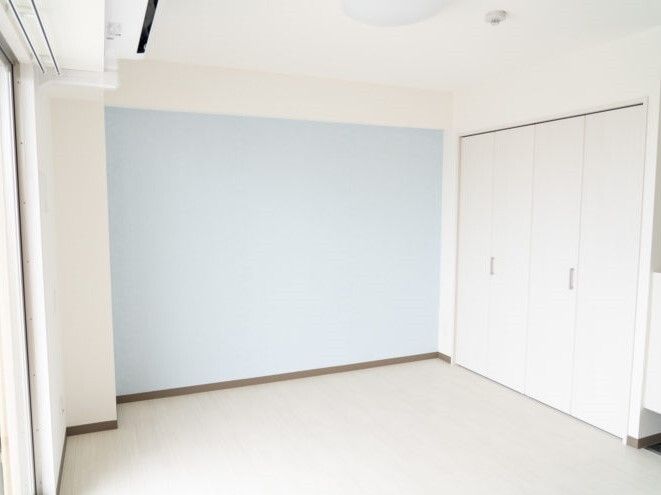 白を基調とした部屋は広く、壁の一面がライトブルー出清潔感がある。右側には収納スペースがあり、左の窓の上の壁にはエアコンが付いている。