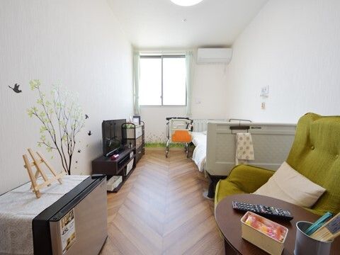 一人用のグリーンのソファやテレビ、寝具付きのベッドやエアコンなどが設置されたフローリングの部屋。壁には木と小鳥のウォールステッカーが貼られている。