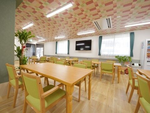 自動販売機や大型の壁掛けテレビが設置された明るい雰囲気の食堂。木製のダイニングテーブルが並び、グリーンの椅子がセットされている。