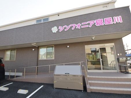 ブラウンと白の2階建ての建物は明るい雰囲気でピンクの文字でホーム名も書かれている。敷地内に駐車場もあり、玄関には階段とスロープがある。