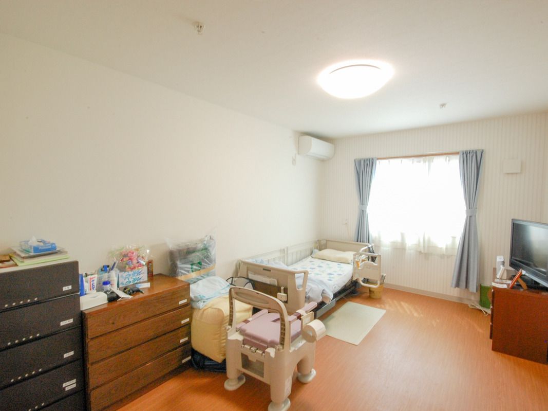 寝具付きのベッドやテレビ、タンス、ポータブルトイレなどが置かれた部屋には大きな窓があり、カーテンも付いている。