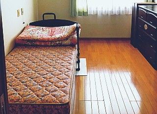 ピンクのエプロンを付けた若い女性スタッフが車いすの高齢女性と日当たりのいい屋内で笑顔で話している。