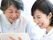 レースの襟が付いたブラウスを着た高齢女性と若い女性スタッフが楽しそうに話をしている様子。二人はスタッフの持っているカルテのようなものを見ている。