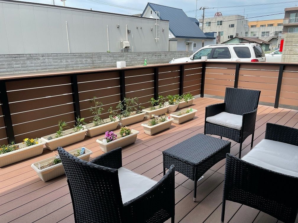 ウッドデッキが敷かれたテラスには何もなく、周辺には建物が多くある。手すりが付いた廊下には大きなガラス戸があり、テラスと行き来ができる。